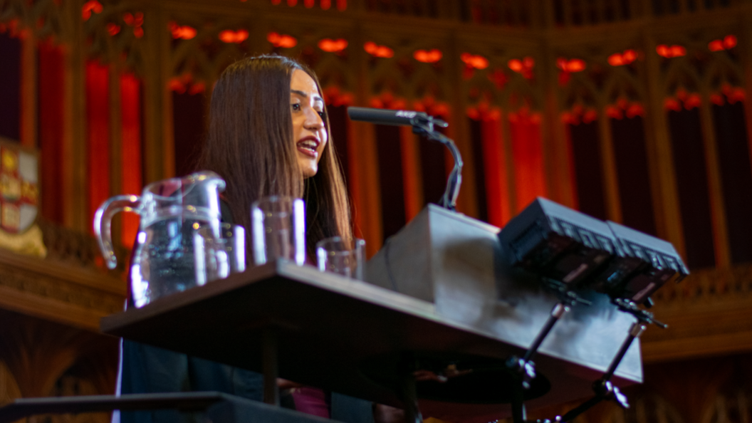 Bristol University masters student Gurvin Chopra is standing behind a lectern speaking into a microphone. She has long brown hair.