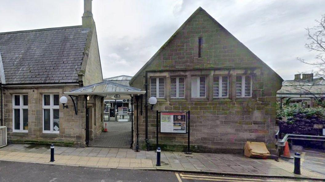 The entrance of platform 2 at Durham train station. Two beige stone building house the station cafe, waiting room and toilets. The glass entry barriers are in the distance. 