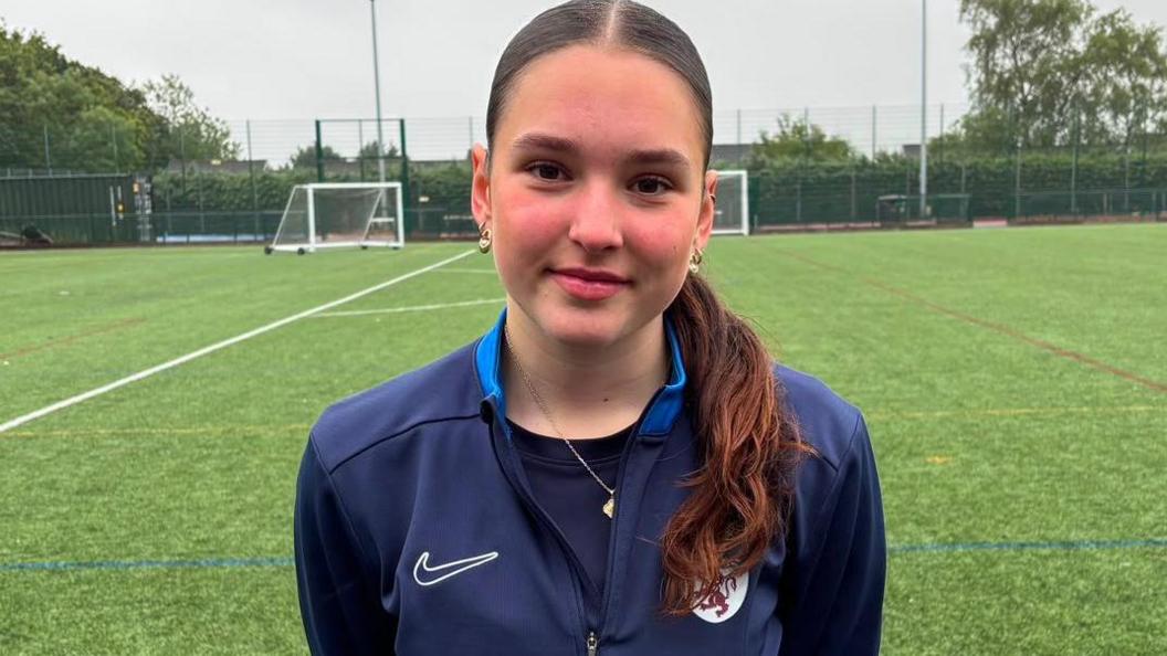 Molly Stroud looks at the camera while standing on an artificial training pitch with goals and fencing in the background