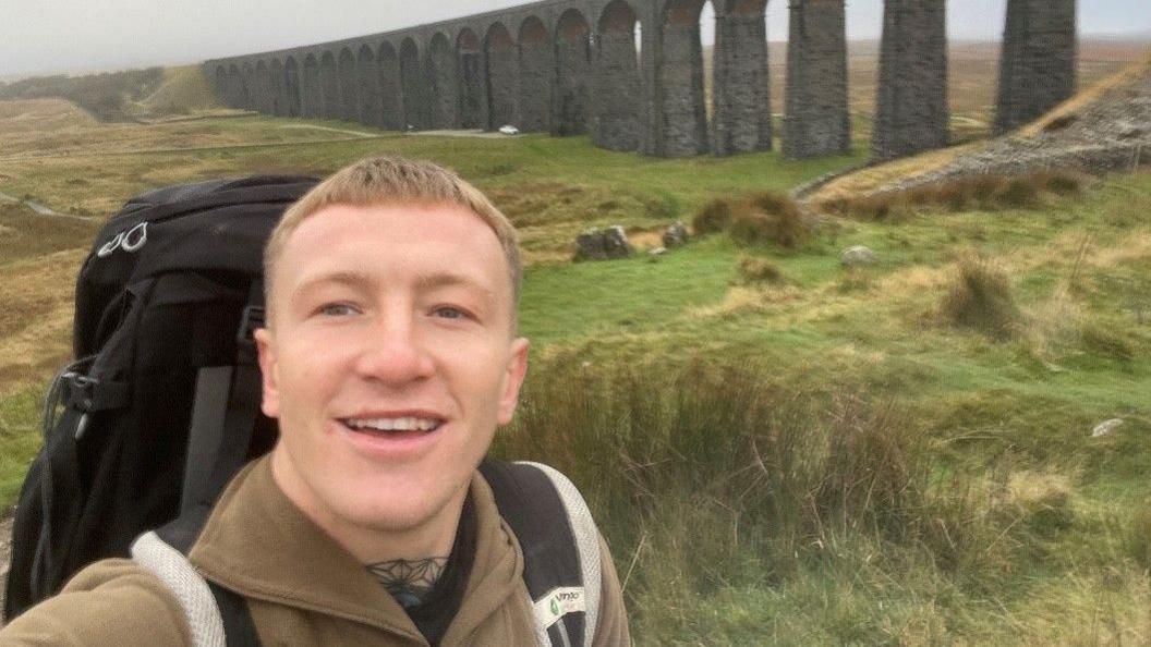 A selfie of man with short blonde hair wearing a brown fleece and large black rucksack smiles with Ribblehead Viaduct in the background on a grey day