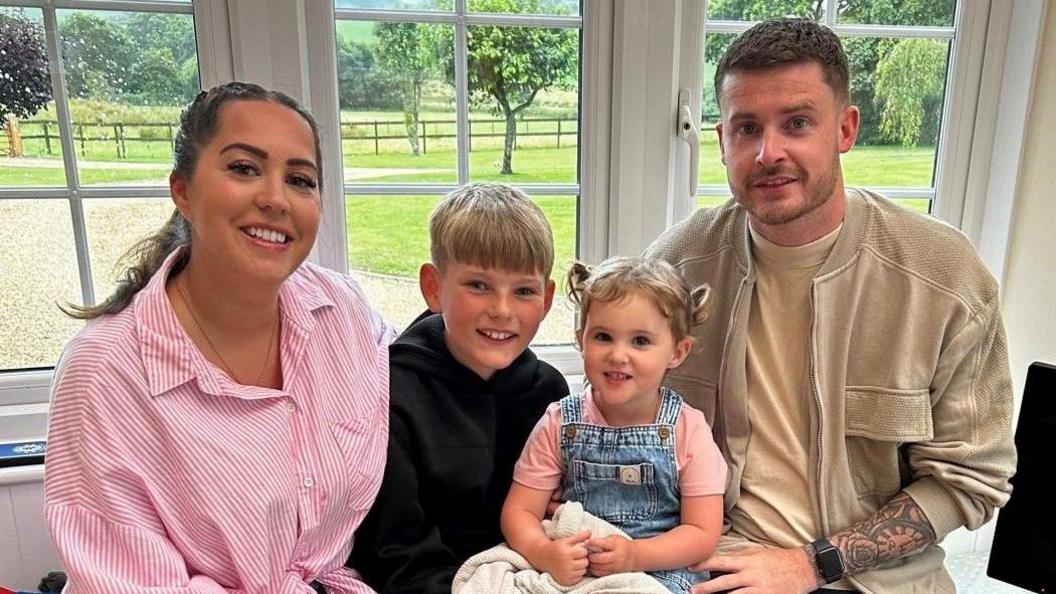 Nikki Roberts sitting down in a window seat with her son Fin, her daughter Cleo and partner Liam. They are all looking at the camera and smiling. Nikki has brunette hair and is wearing a white and pink striped shirt. Fin has short blonde hair and is wearing a black hoodie. Cleo has two little pony tails in her blonde hair and is wearing denim dungarees and a pink T-shirt. Liam has short blonde hair and is wearing a beige jacket and yellow T-shirt. He has a tattoo and a smartwatch on his arm.