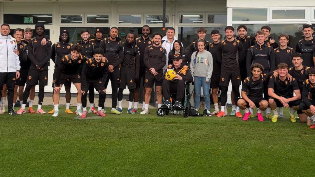 The Hull City squad in matching kits. They're stood on the grass pitch in front of the windows with Sophie, her partner and her dad all stood beside them