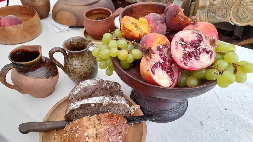 A display of food on a white tablecloth. A dark wood fruit bowl displays green grapes, pink pomegranates with red seeds, and dark pink peaches. There is slices bread on a wooden plate, and ceramic milk jugs