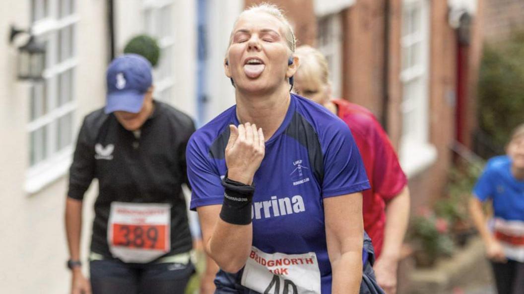 A female runner signals her tiredness on a steep hill by pulling a face with her tongue hanging out. The woman has blonde hair and is wearing a purple t-shirt, sweatbands on her wrists and earphones. A running number is just visible pinned to her t-shirt, saying Bridgnorth 10k. Other female runners are visible out of focus behind her.