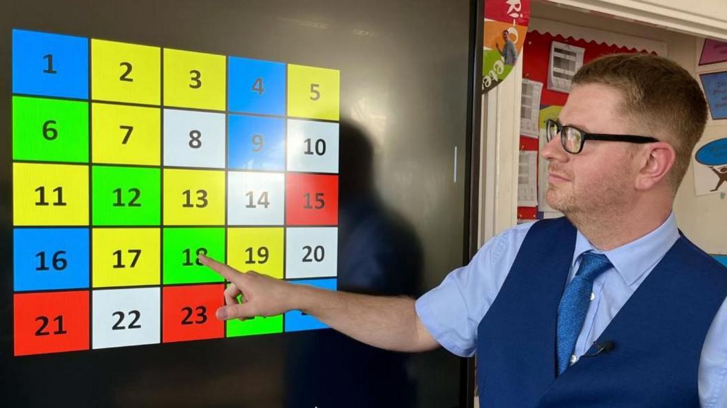 Norfolk teacher Tom Chapman pointing at an interactive classroom board featuring a five by five grid of numbers on different coloured backgrounds