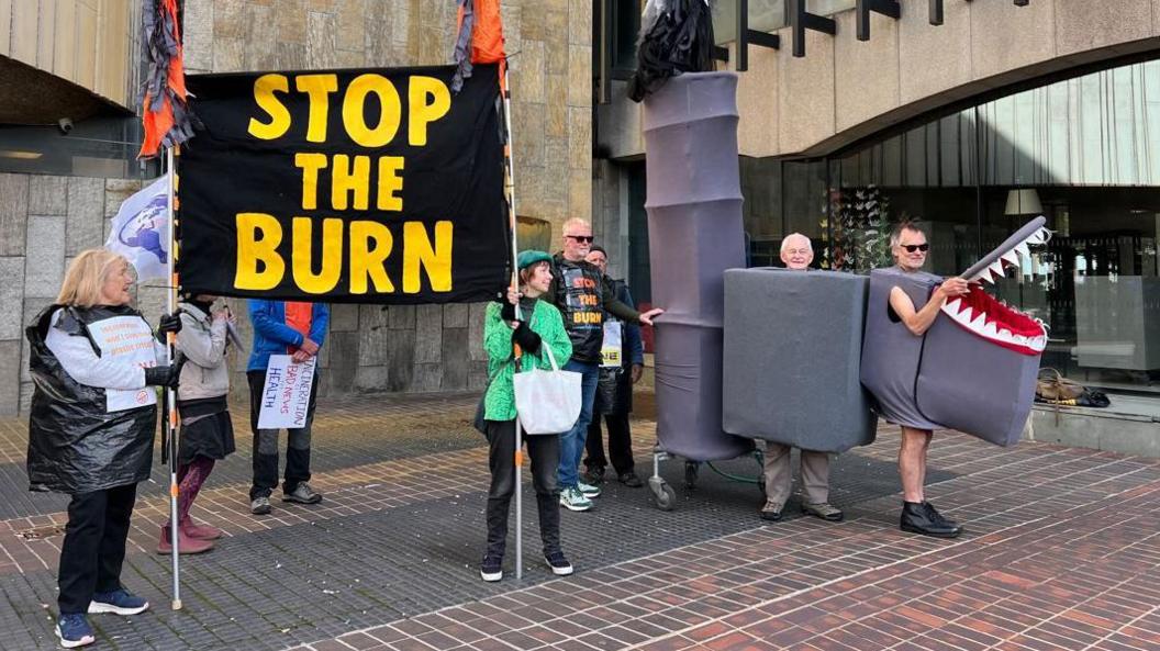 A group of seven people, one in a bin liner holding one side of a banner stating "stop the burn" with a woman in a green jacket holding the other. Two men are dressed as grey incinerator, one in a box, with the other in another box with a representation of sharks teeth around the edges of its open lid