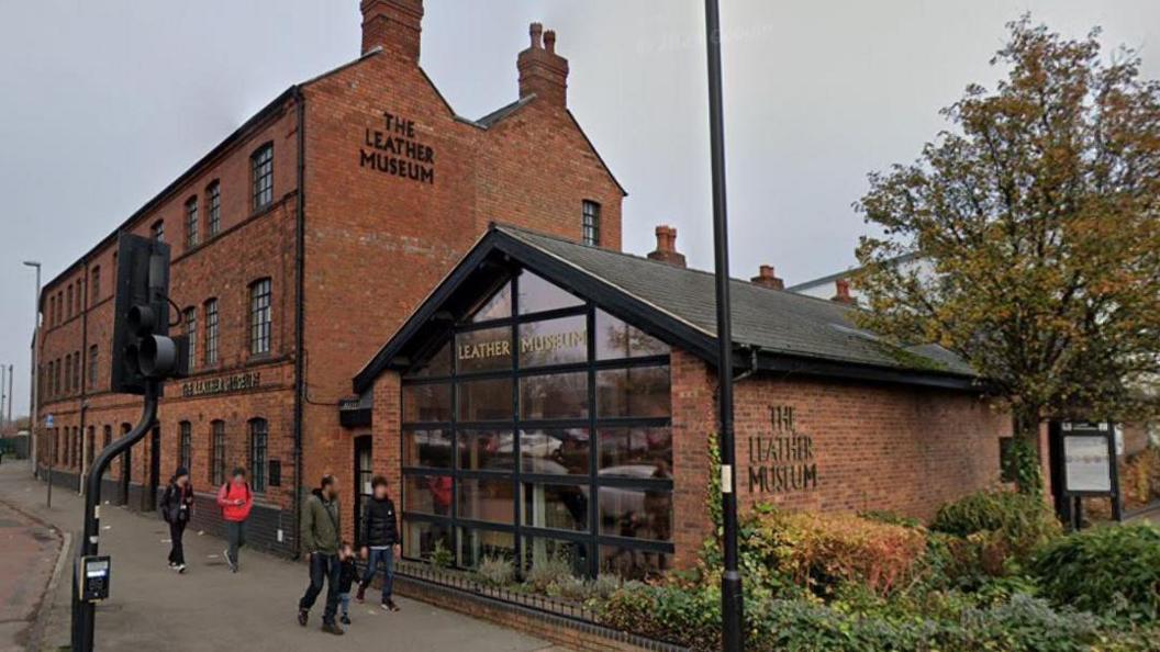 A large red brick building with black framed windows and the words The Leather Museum in black lettering on the walls. There is a glass fronted building in the foreground and people walking past.