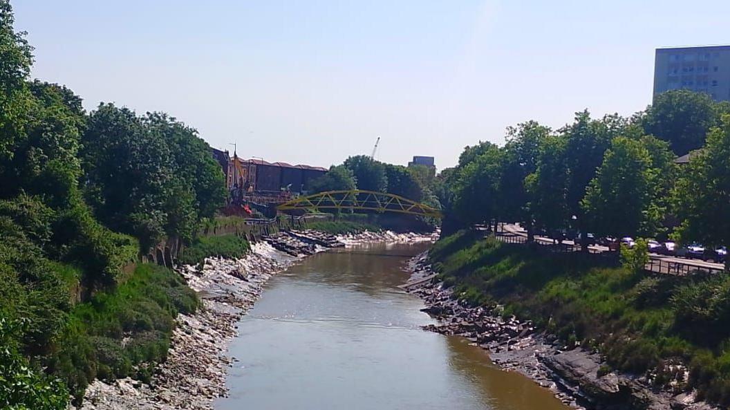 The Avon New Cut near Temple Meads station on a sunny day, with the yellow "banana bridge" crossing the river and trees lining the river.