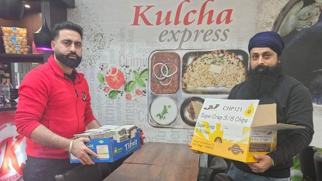 Soul Aid Founder Deepak Singh and Owner of Kulcha Express, Ramjinder Maan Palvinder Singh each carrying a box of free food and standing in front of a colourful poster with pictures of rice and curry.