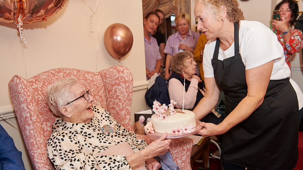 Annie is seated and being presented with a cake by a staff member in a black apron and white top