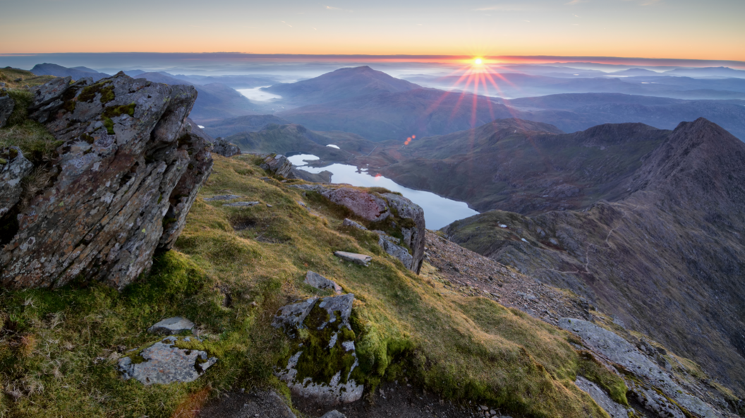 A vast, mountainous landscape is pictured from a summit looking down at lakes, clouds and grassland at sunrise.