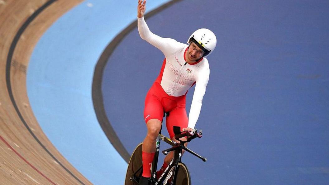 Dan Bigham celebrates after the Men's 4000m Individual Pursuit qualifying at Lee Valley VeloPark on day two of the 2022 Commonwealth Games in London. He wears a tight-fitting white and red outfit and waves an arm while riding on a bike