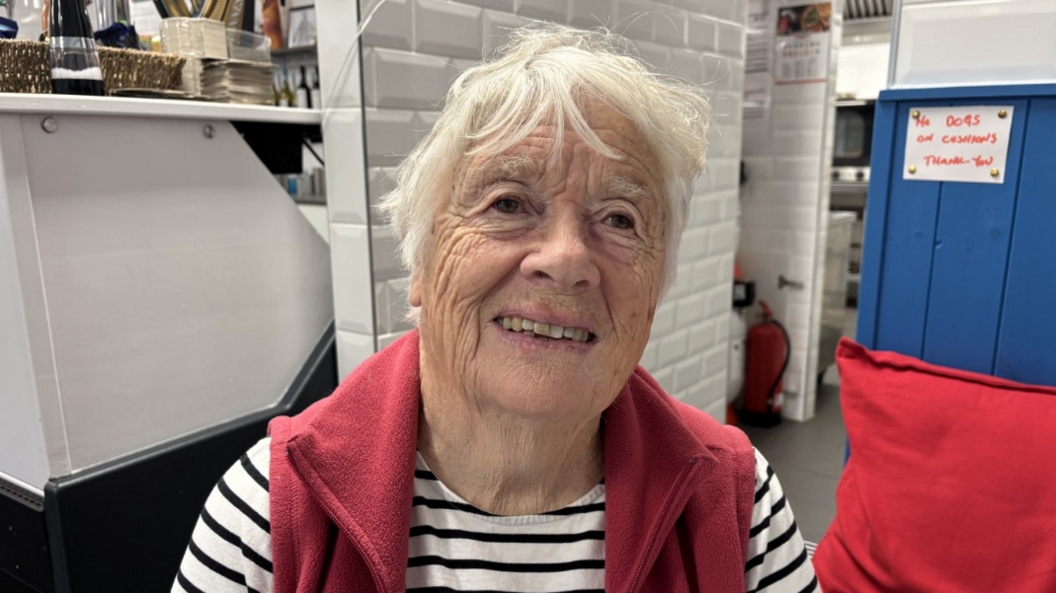 Close up of a woman smiling at  camera. She is wearing a black and white striped top and a red gilet. She is sitting inside a cafe.