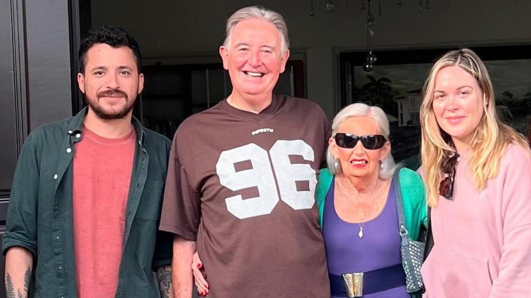 Four people are stood next to each other. From left to right - a man with beard and moustache and dark hair is wearing a red t-shirt and green shirt. Next to him is an older man, wearing a brown t-shirt with the number 96 on it. Next to him is an older woman with long white hair, wearing sunglasses. She is also wearing a purple top with belt and green cardigan. At the end is a young woman with long blonde hair and pink top with sunglasses hanging from the collar.