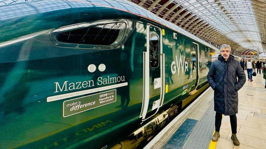 A man with grey hair is wearing a coat on the right hand side of the photo. The image is dominated by a green train at a railway station. On the left of the image the train has the words Mazen Salmou on it.