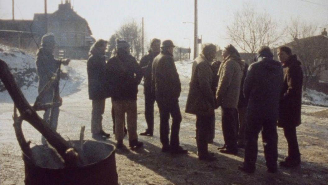 A group of men standing on the road outside the entrance to Oakdale pit. It's winter and there's some snow on the ground. The men are wearing large coats and some are wearing hats and gloves. In the foreground there's an empty oil drum with smoking tree branches sticking out of the top. The background there are some bare trees and the start of a row of terraced houses.