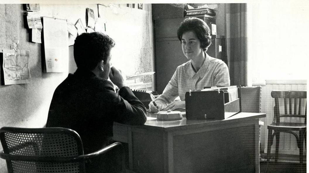 Black and white photo shows two people sitting on opposite sides of a desk. One, whose back is to us, has a suitcase, is opposite a woman in a light shirt
