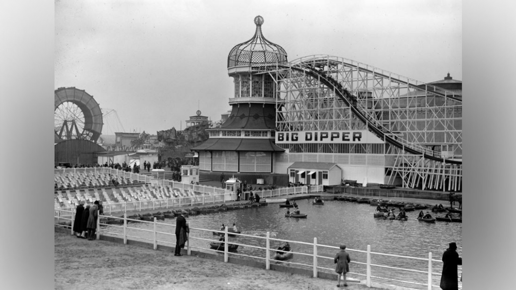 A tall rollercoaster stands in the foreground with a lake in front of it with people on boats, a Ferris wheel can be seen to the left of the rollercoaster along with a deck chair seating area.