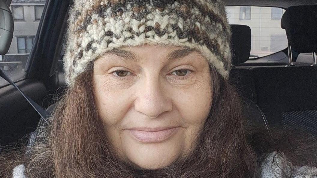 A middle-aged white woman sitting in a car and taking a selfie. She has dark hair and is wearing a knitted hat. There are houses in the distance.