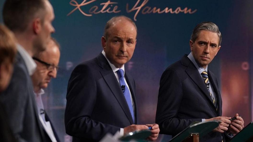 Micheál Martin and leader Simon Harris look to their right as another party leader speaks during a debate. Both men have dark suits on and are holding a pen in their hands 