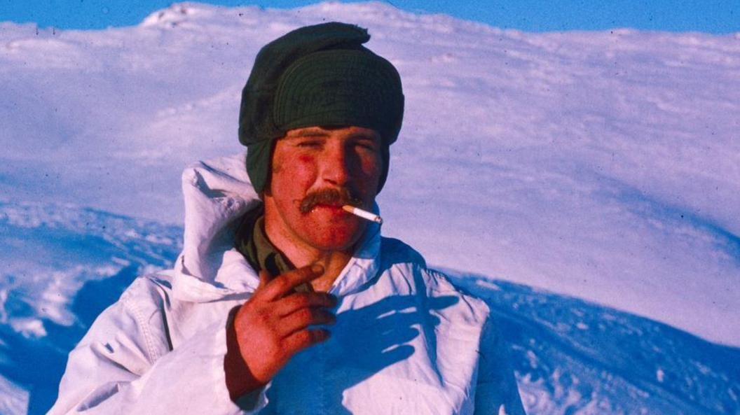 Rob Ewen stood in the snow with a black hat and a white coat. He has a cigarette in his mouth and of course, has a large moustache. He is looking at the camera and smiling. 