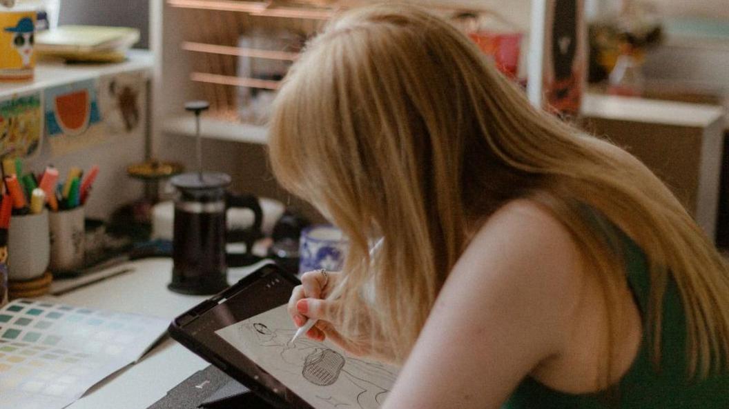 Ana sits at a desk which has stationary placed on it. She is working on an illustration on an iPad.