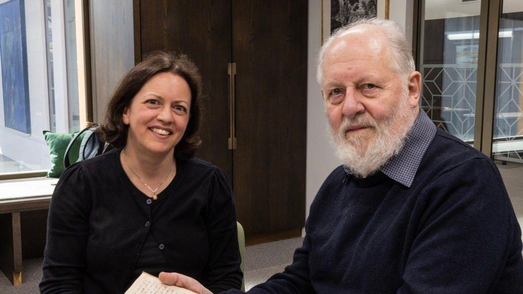 A woman with a dark brown bob wearing a black cardigan and a man with short grey hair and bear wearing a navy jumper and shirt.