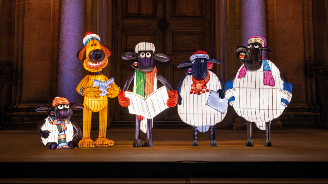 Illuminated lantern sculptures of Shaun The Sheep and other sheep friends - and a dog - standing in a darkened doorway of Longleat House. They are wearing Christmas hats and scarves, holding booklets as if they are singing Christmas carols.