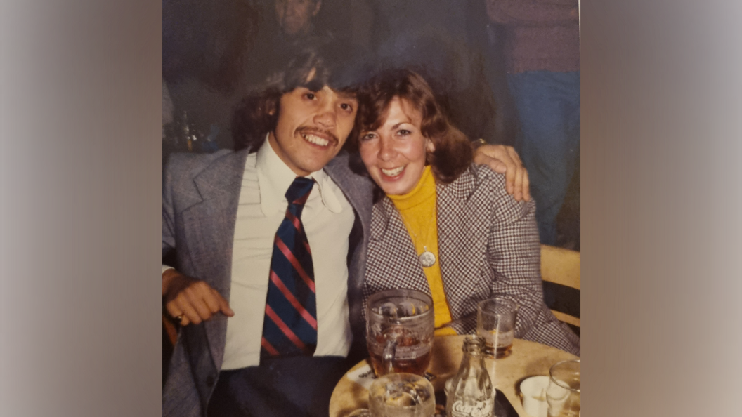 A young man wearing a suit, smiles at the camera, as he hugs his wife, who is also smiling at the camera, wearing a yellow top and checked blazer as they sit in a pub.