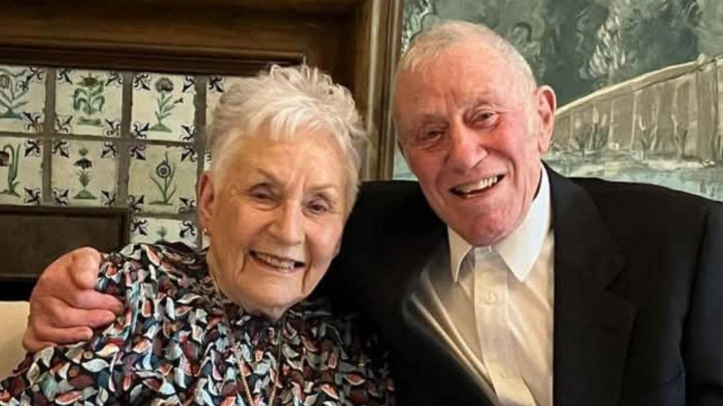 Veronica Ash and her husband are smiling for the camera. Ms Ash is wearing a patterned top and her husband is wearing a black suit jacket and a white shirt.