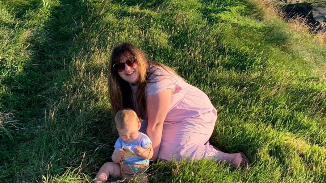 Amy and her baby are pictured sitting in a field on a sunny day. She has long dark hair and is wearing sunglasses, the baby is wearing a short-sleeve top and his legs are bare.