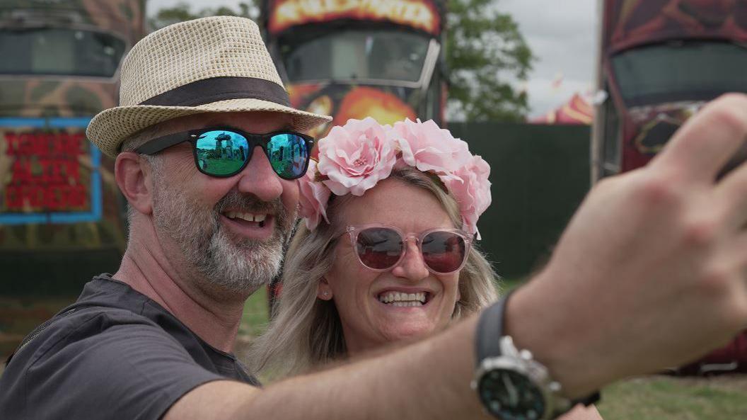 Man in hat and woman with flower hairband smile whilst taking a photo together