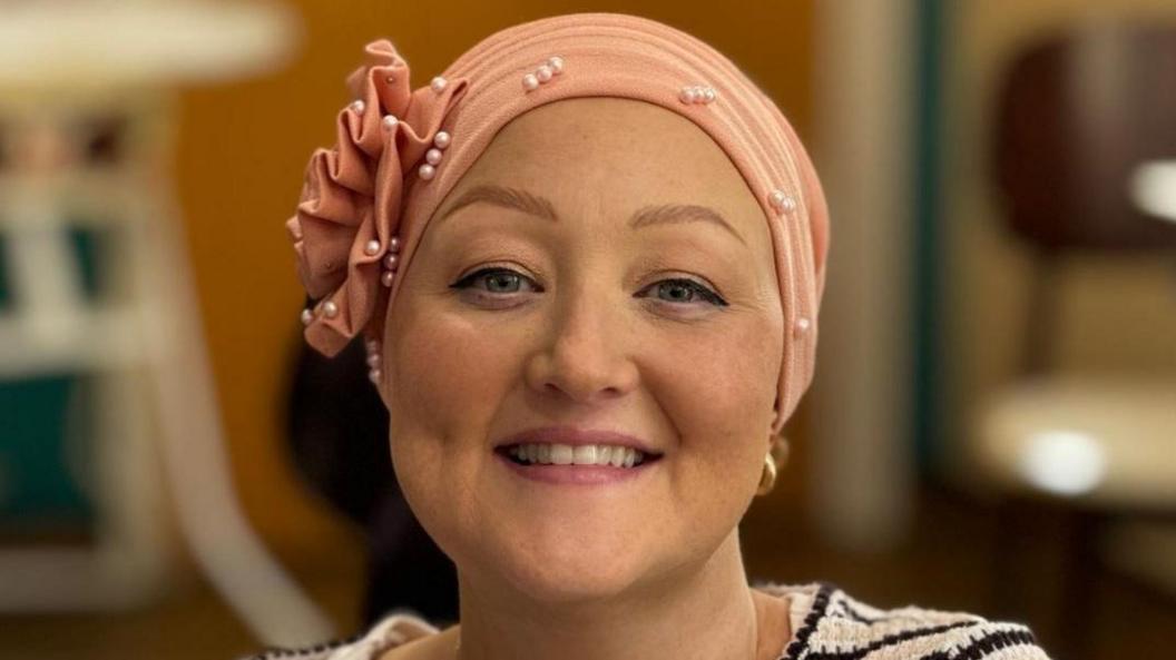 A woman smiling at the camera in a hospital room. She has blue eyes and is wearing make up and a pink headscarf decorated with a ruffle and pearls. She is wearing a striped top.