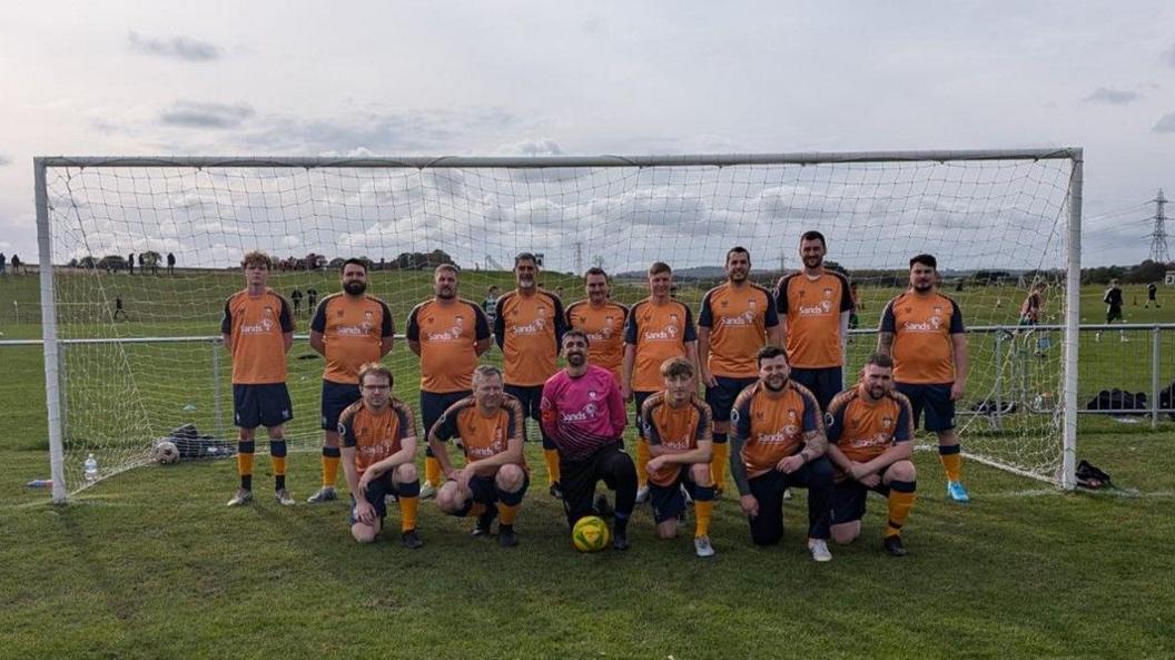 16 men on a green football pitch in front of a white net. 15 Men are wearing orange and black t-shirts and shorts while there is a man, the goal keeper, wearing a long sleeve pink shirt and black jogging bottoms.