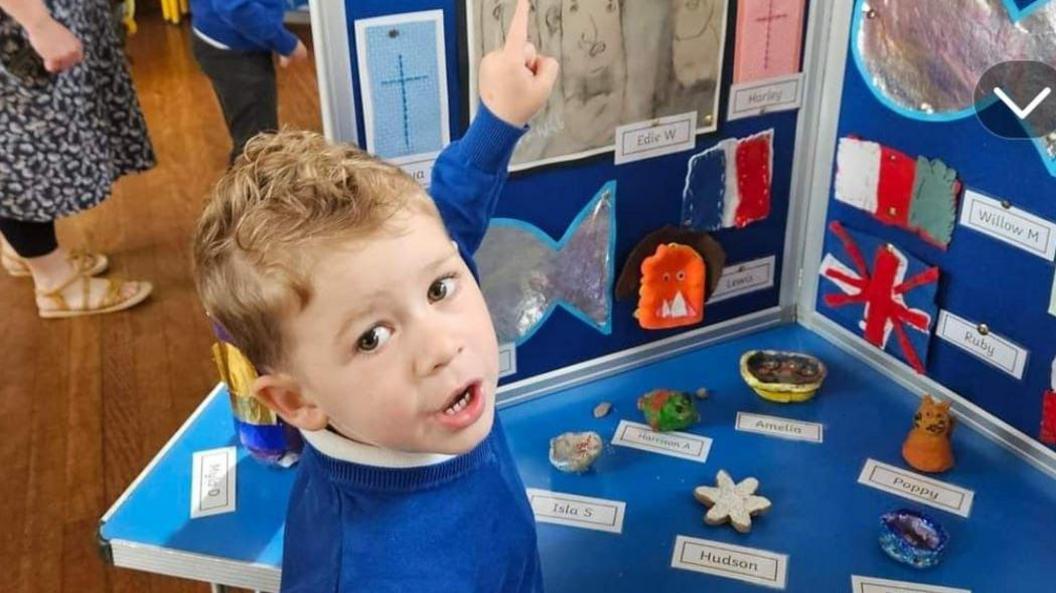 A little boy in a blue school jumper points at an art display.