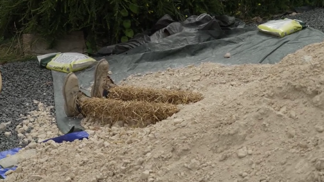 A pile of sand next to a construction site has had two straw legs, with shoes on, added to the end of it. It looks as though a scarecrow is stuck under the sand.