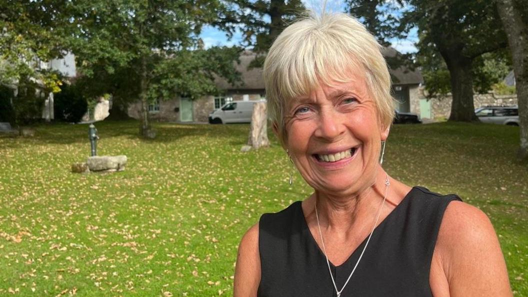 Sally Everton from Visit Devon wearing a black sleeveless dress and a silver necklace. She is standing on the gree in North Bovey with an old water fountain in the background to the left of frame.