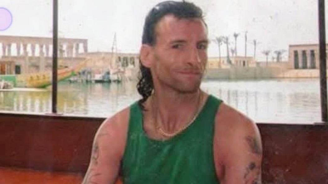 Timmy sat on a boat looking at the camera with a slight grin. He is wearing a green vest top and a chain necklace. He has a dark brown mullet and has a few tattoos on his arms.