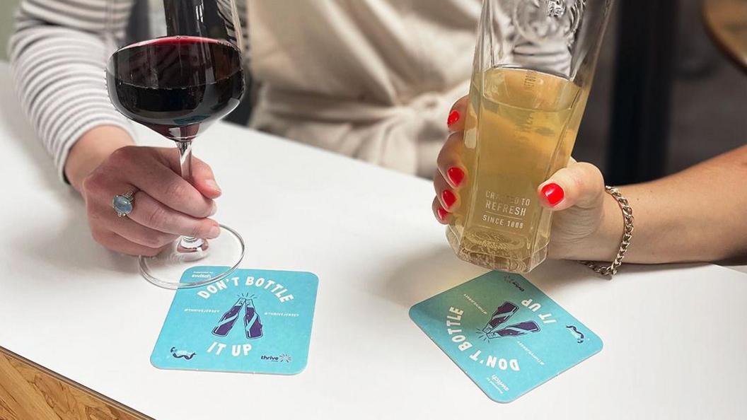 The arms of two women, one with a striped long shirt and ring on her middle finger holding a large red glass of wine, and the other with a gold chain bracelet on and red nails holding a cider in a pint glass, with two blue beer mats below the glasses with the campaign slogan 'Don't bottle it up' with two beers clinking in the middle on a white and brown oak table