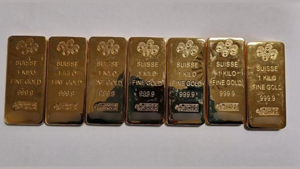 Seven gold bars in a row on a light grey surface. All of the bars have the words 'Suisse', '1 kilo', 'fine gold' and '999.9' printed on them alongside other markings.