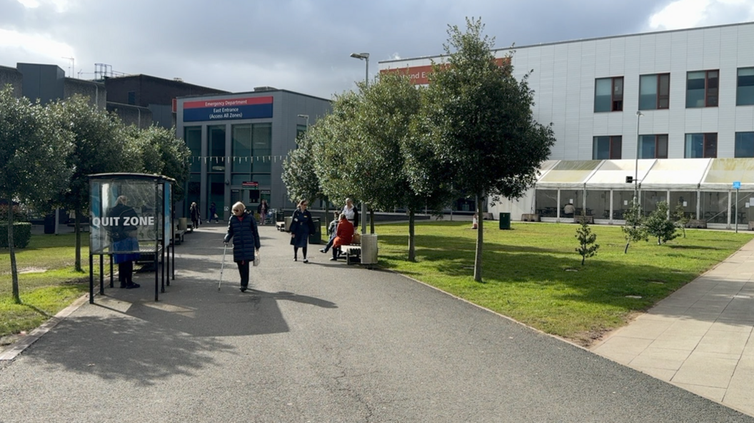 The entrance to the hospital has some trees along with path towards it with a small smoking hut on the left. A white building can be seen on the right.