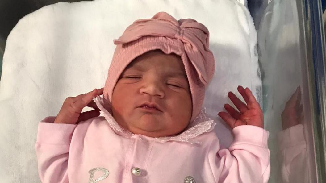 A newborn baby girl wearing a pink hat and pink overalls. 