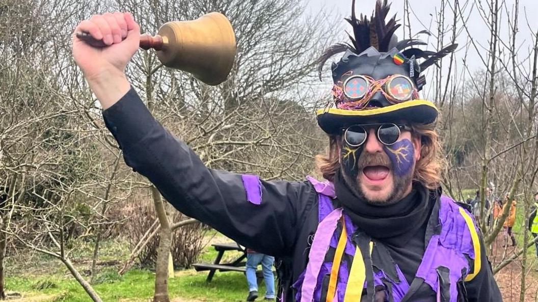 Jamie wears a top hat with feathers sticking out the top and a coat with purple, orange and yellow ribbons hanging from it.  He wears purple face make up over a beard and black sunglasses.