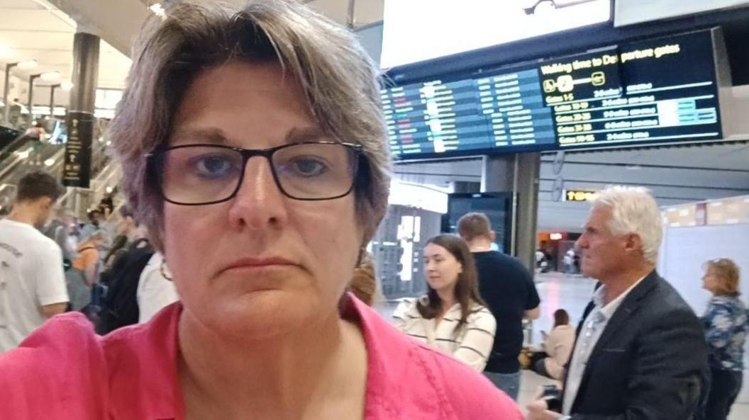 Jayne Ozanne, wearing glasses and a pink top, looking unhappy in an airport departures area