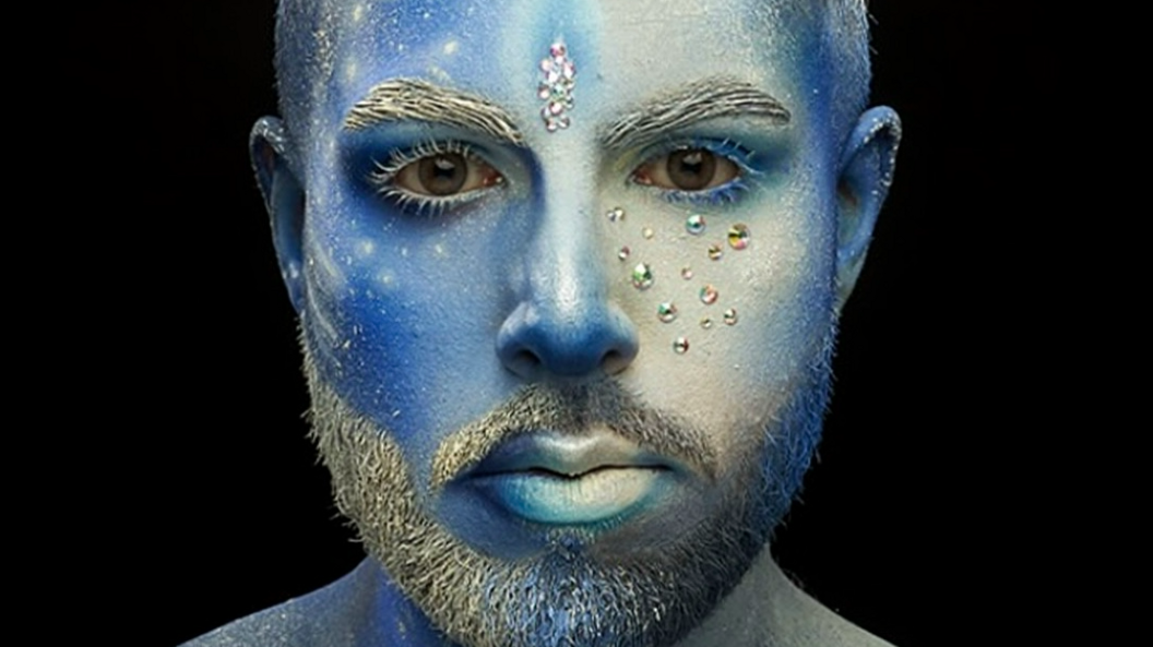 Rahual has a full-face make-up look and is looking into the camera. His entire face and shoulders have been painted in different hues of blue. There are white highlights and accents, such as paint splatters on one side of the face that look like a cluster of stars. His eyelashes, eyebrows and beard are painted white. In between his brows is a sparkly bindi.