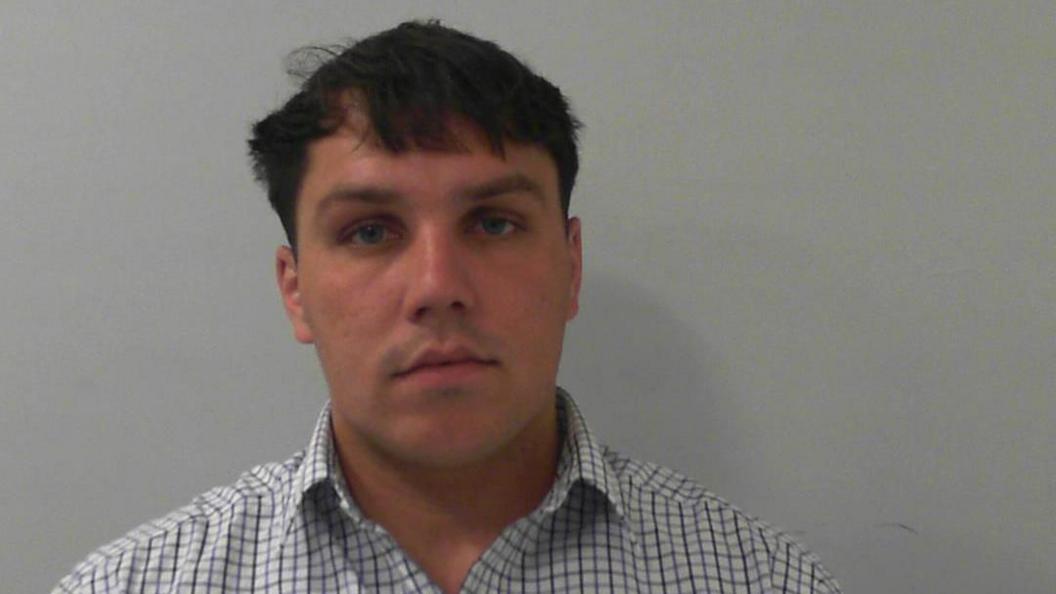 A police mugshot of Archie Stevenson, who has mid-length dark hair and is wearing a chequered shirt.