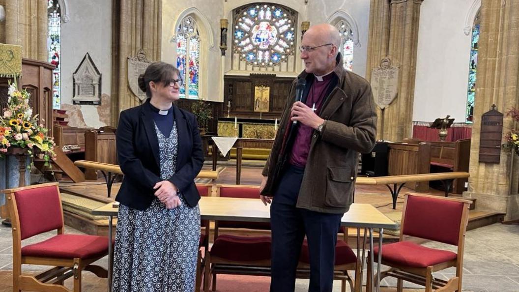 Dr Fiona Gibson wears a blue floral dress black jacket and dog collar looks at Bishop Michael Beasley. He is wearing a wax jacket, pink shirt, dark trousers and dog collar. They are standing in front of an alter in an ornate church with stained glass windows. The man is holding a microphone.