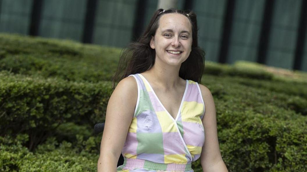 A woman with long brown hair is looking at the camera and smiling. She is visible from the waist up, and is wearing a multicoloured pastel vest top with green, yellow, purple and pink squares. She is in front of some hedges in the background.