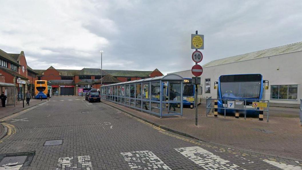 A Google Streetview of the bus station in Carlisle. It is a wide cobbled dead-end street with different bays with bus shelters on one side and parking bays for buses on the other side. There are some shops at the bottom of the road.