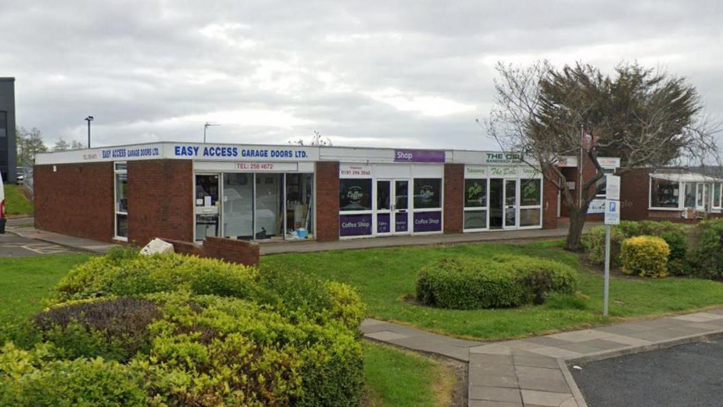 A row of three business units next to a footpath and grass. The left one is East Access garage Doors, in the middle is a vacant cafe and to the right is The Deli.